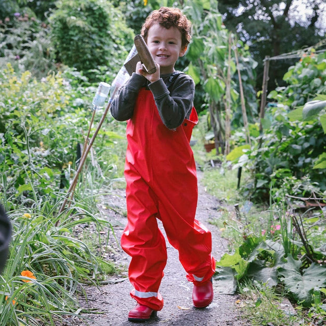 A child in red overalls & wellies walks along a path in a lush garden, carrying a gardening tool with a big smile.